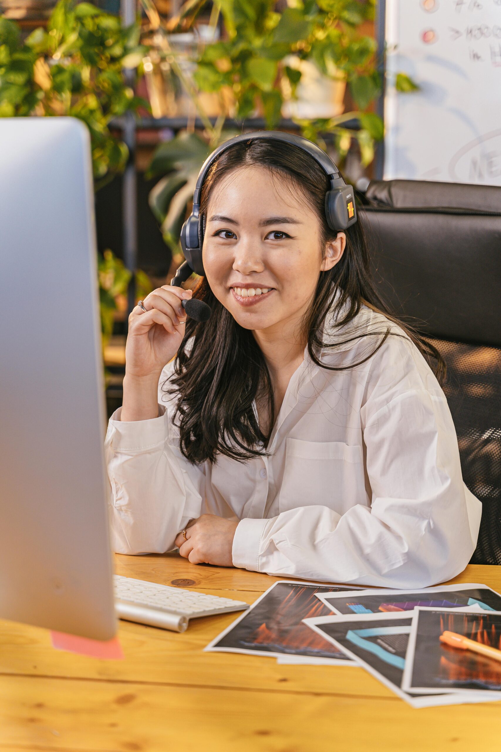 Asian woman smiling while working at a desk with a headset and computer indoors.